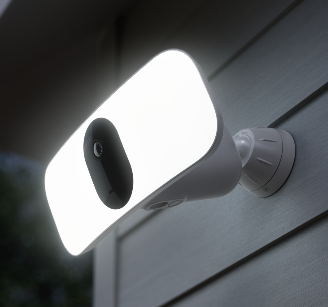 Arlo Floodlight, zewnętrzna kamera bezpieczeństwa z wbudowanym reflektorem o dużej mocy, zainstalowana przy ścianie w nocy, oświetla otoczenie mocą reflektora. 